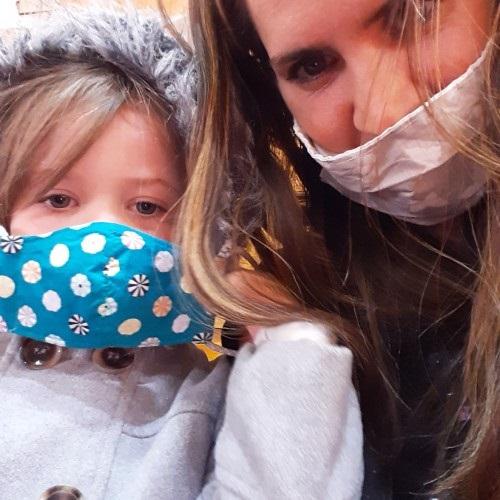 A mom and her daughter looking down at the camera. The mom is wearing a beige fabric face mask with white butterflies. The daughter is wearing a blue fabric face mask with flowers.