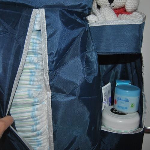 There is a hand opening the nappy pocket of the nursery organizer. The pockets on the side are filled with baby items such as creams and knitted teddy bear.